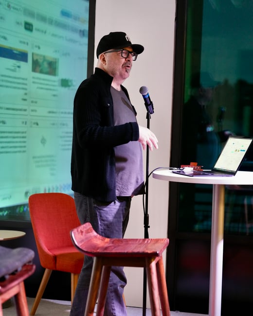 A man wearing a black cap, glasses, and a dark cardigan stands next to a microphone, giving a presentation in front of a large projected screen displaying web content or a dashboard.