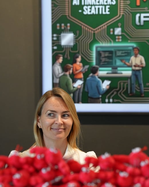 A portrait of a smiling woman standing behind a blurred pile of red plush toys, with a screen in the background displaying an 'AI Tinkerers - Seattle' graphic.