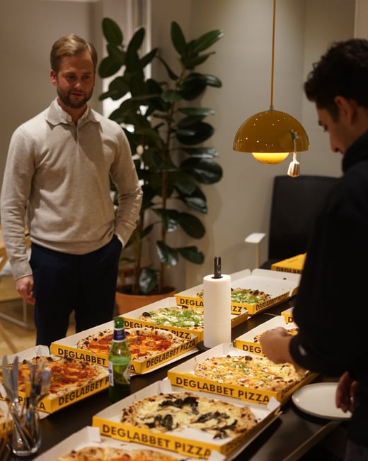 A candid indoor photo showing two men standing near a long table laden with multiple open pizza boxes from "Deglabbet Pizza," likely during a casual social gathering or meal.