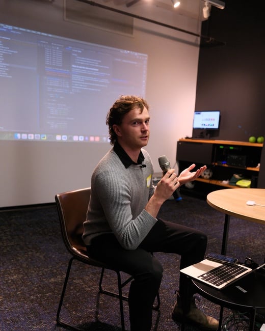 A man wearing a gray sweater is seated and speaking into a microphone during a technical presentation or Q&A session in a dimly lit indoor venue. Behind him, a large projection screen displays lines of code or software interface elements.