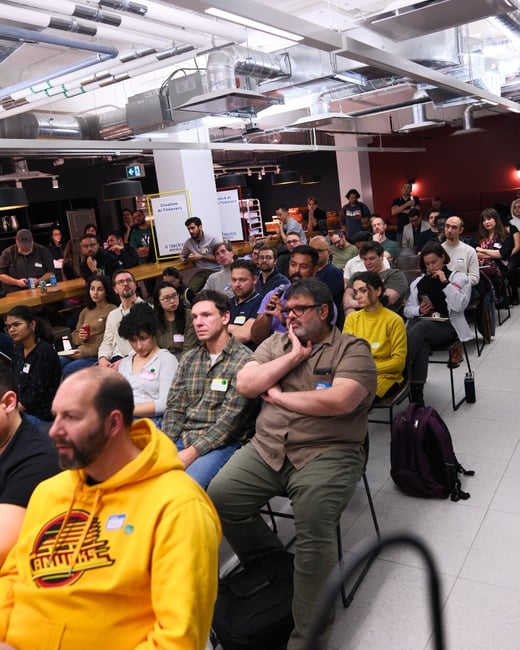 A candid photograph capturing a large audience seated indoors, attending an event or presentation in a modern space characterized by high ceilings and exposed industrial ductwork.