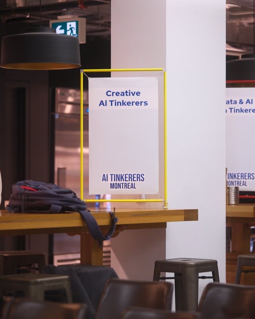 A photograph capturing an indoor event or co-working space, featuring a high wooden table, dark stools, and a sign framed in bright yellow advertising "Creative AI Tinkerers" and "AI Tinkerers Montreal."