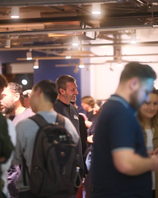 A candid photograph capturing a group of people socializing indoors at an event, featuring a smiling man with a beard in the center-left middle ground and a person wearing a dark backpack blurred in the immediate foreground.