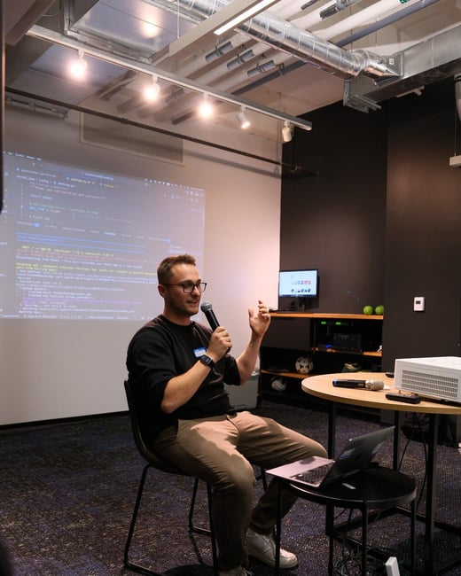 A man wearing glasses and a black shirt is seated, holding a microphone, and giving a technical presentation in an indoor space with exposed industrial ceilings.