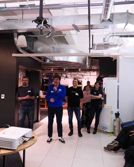 A photograph captures a group of seven people gathered indoors, likely during a tech event or presentation, where one man in a blue sweater is speaking into a microphone. The setting features an industrial-style ceiling with exposed ductwork and a white projector visible on a small table in the foreground.
