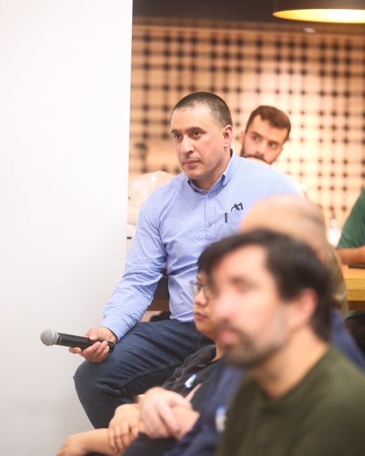 A candid indoor photo capturing a man in a blue, polka-dot shirt holding a microphone while seated among other attendees at an event or meeting. The man is in focus, while the foreground figures and background details, including a patterned wall, are softly blurred.