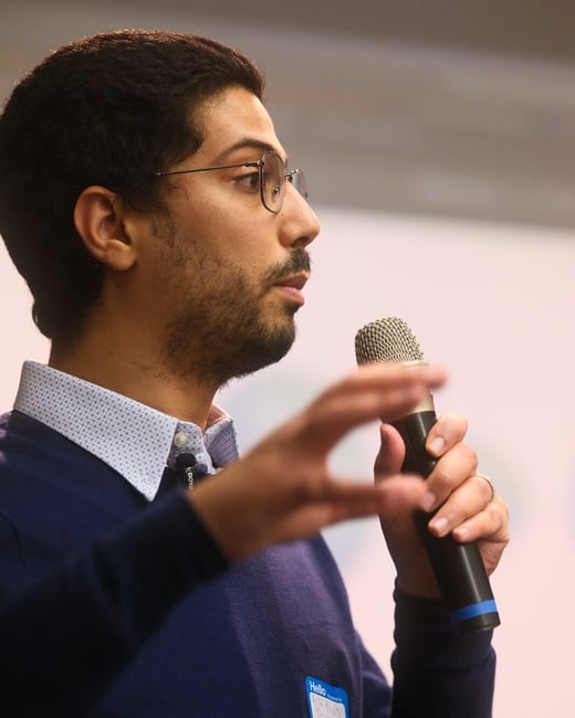 A close-up, profile photograph captures a man wearing glasses and a dark blue sweater speaking into a handheld microphone, likely during a presentation or public event.