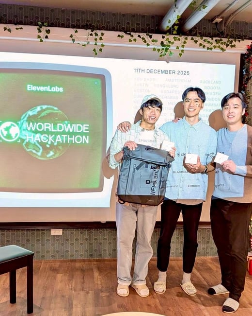 Three young men stand together and smile in front of a large projection screen at an indoor event, holding prizes including a gray backpack and small white boxes. The screen displays information for a "WORLDWIDE HACKATHON" hosted by ElevenLabs, along with a date and a list of global cities.