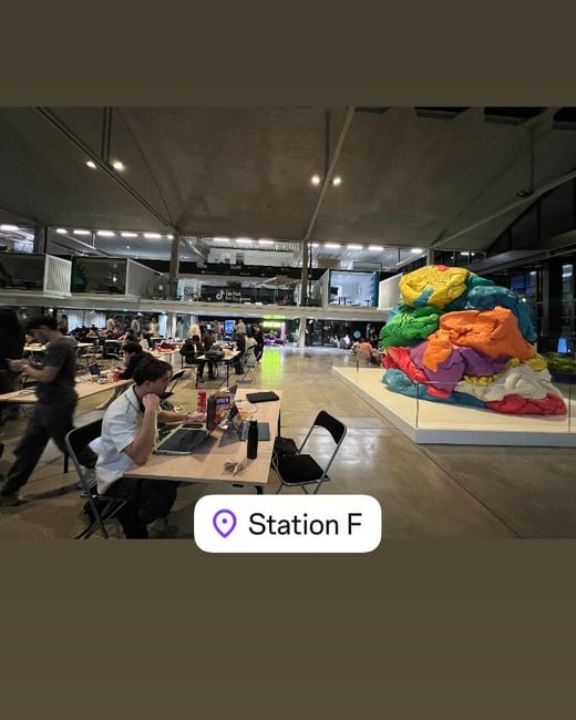 A candid photograph taken inside a large, industrial-style co-working or event space, likely Station F, showing many people seated at tables working on laptops, with a large, brightly colored abstract sculpture displayed prominently in the center-right background.