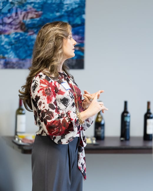 A woman in a floral blouse and grey trousers stands in profile, gesturing with her hands in an indoor setting with wine bottles in the background.