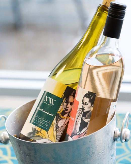Two bottles of wine, a Chardonnay and a Rosé, are chilling in a silver ice bucket. The bottles feature artistic labels with illustrations of women.