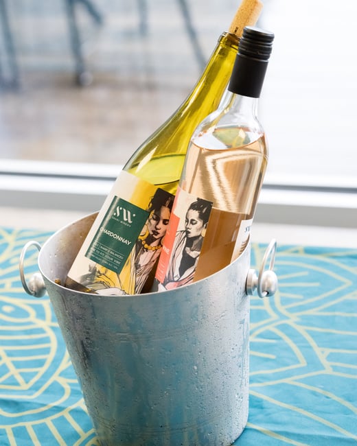 Two bottles of wine, a Chardonnay and a rosé, are chilling in a silver ice bucket on a blue patterned tablecloth.