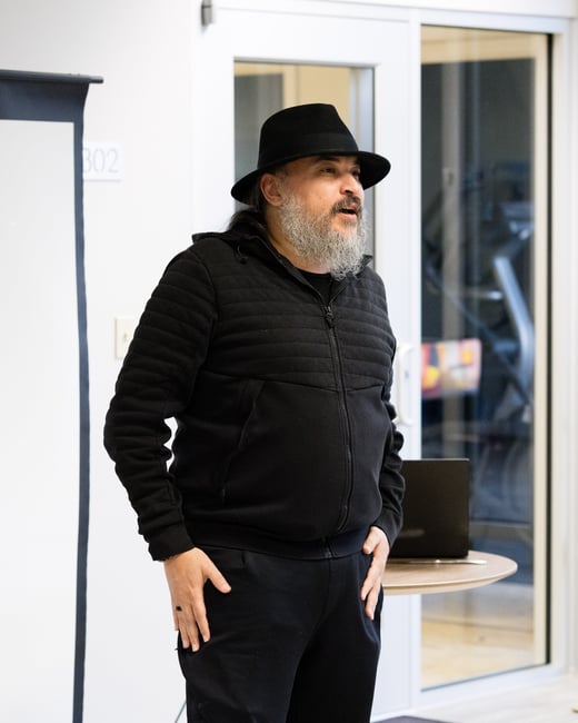 A man with a grey beard wearing a black hat and jacket is standing and speaking in an indoor setting, likely giving a presentation.