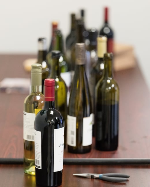 A collection of various red and white wine bottles standing on a dark wooden table, with a pair of pliers and a corkscrew in the foreground.