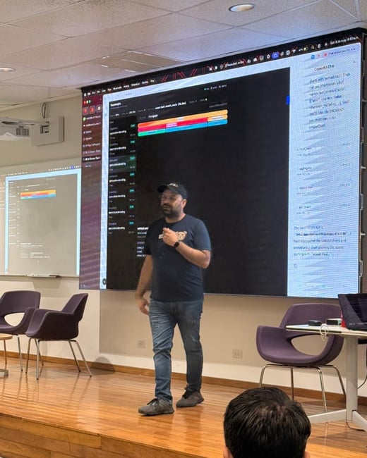 A man stands on a wooden stage, delivering a presentation in front of a large screen displaying a code editor and a chat interface.