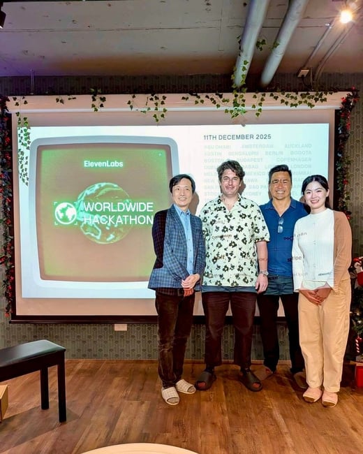 A group photo of four individuals (three men and one woman) standing indoors in front of a large projection screen advertising the ElevenLabs Worldwide Hackathon scheduled for December 11, 2026, listing various global cities.