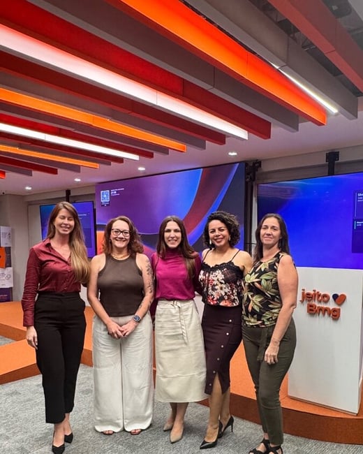 Five women are posing for a group photo in a modern office or event space with distinctive orange ceiling slats and large digital screens in the background.