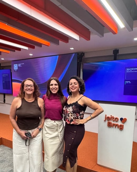 Three women are smiling and posing for a photo in a modern corporate event space featuring a podium and digital screens.