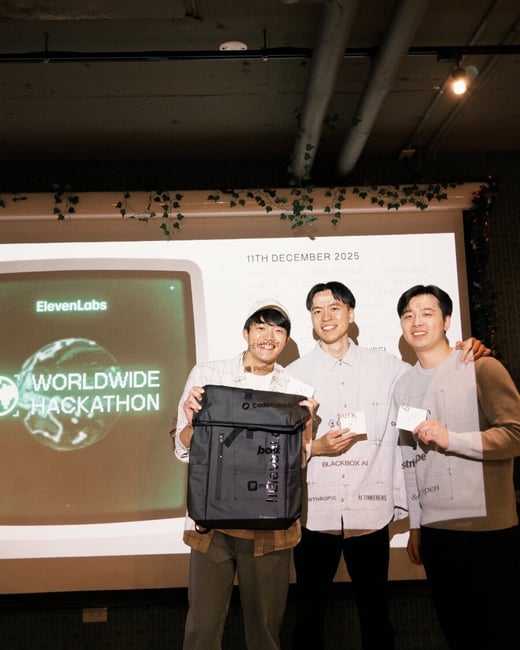 A low-light photograph captures three young men smiling and posing in front of a projection screen at an indoor event, likely a hackathon, with the man on the left holding a black backpack prize.