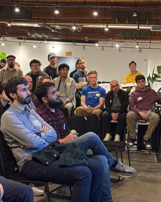 A candid photograph captures a diverse group of people, mostly men, attentively seated and standing in an industrial-style indoor venue, likely attending a presentation or meeting.
