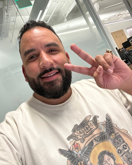 A man with a beard and dark hair is taking a selfie while making a peace sign. He is wearing an off-white graphic t-shirt in what appears to be a modern office setting.