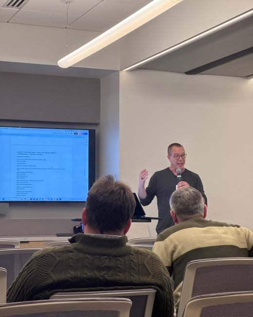 A man is giving a presentation or lecture in a modern conference room, standing next to a large screen displaying tax form data, while two audience members are visible seated in the foreground with their backs to the camera.