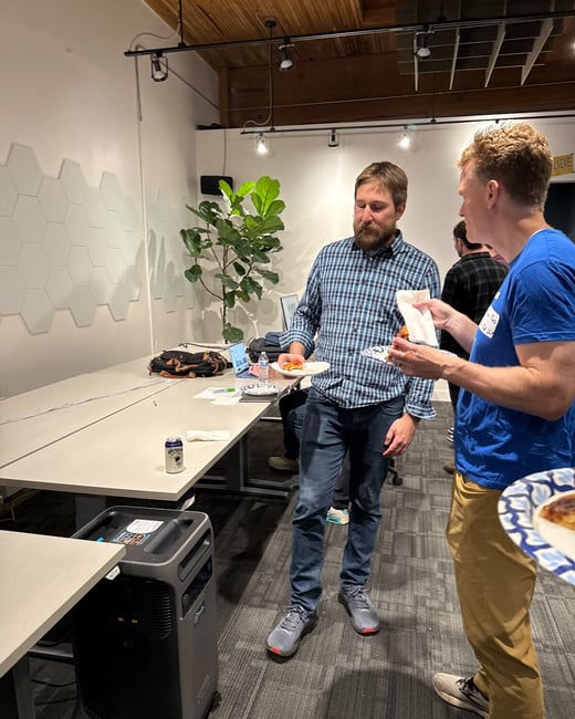 Two men are standing and talking while holding plates of food (likely pizza) and drinks at an informal social gathering inside a modern office or event space.