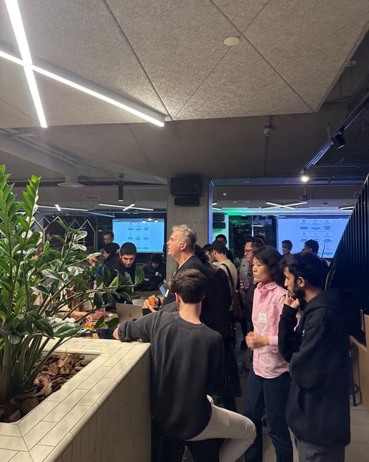 A candid photograph capturing a crowded indoor technical event or social gathering, showing attendees standing and conversing near a tiled counter and a large potted plant, with screens visible in the background.