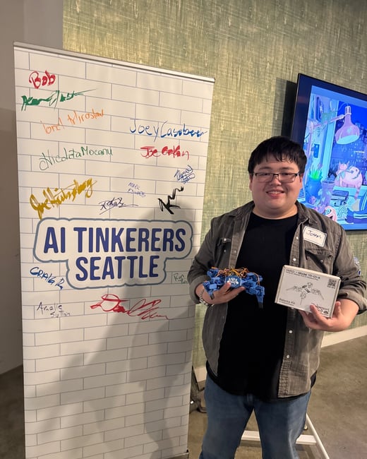 A young man, wearing glasses and a name tag reading 'Joey', stands indoors holding a small blue, wired robot and a white robotics kit box, next to a pull-up banner for 'AI TINKERERS SEATTLE' covered in signatures.