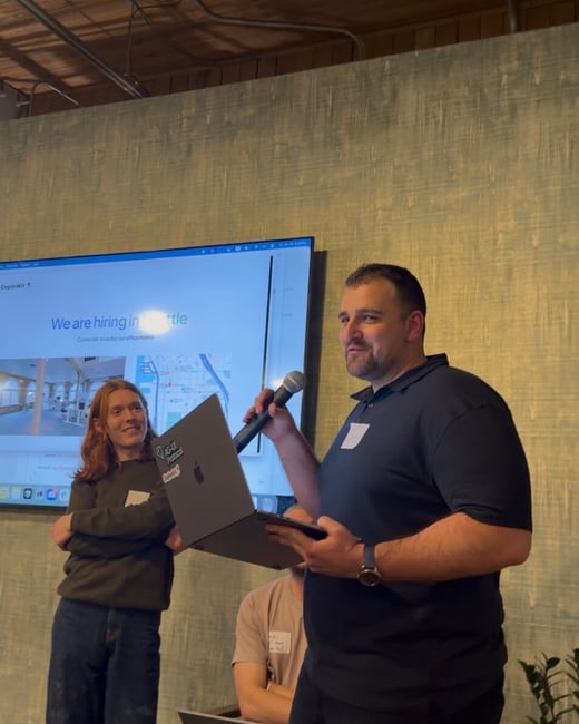 A man speaks into a microphone while holding a laptop during a presentation or event, standing beside a woman in front of a large screen displaying a slide about hiring in a city. The scene takes place indoors with a textured teal wall and an exposed wooden ceiling.