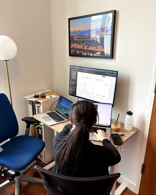 A woman with long dark hair tied in a ponytail is sitting at a desk, working with two laptops and a large external monitor in a home office setting.