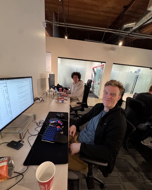 A candid photo taken inside a modern office space, showing two men seated at their desks who are smiling and looking directly at the camera.