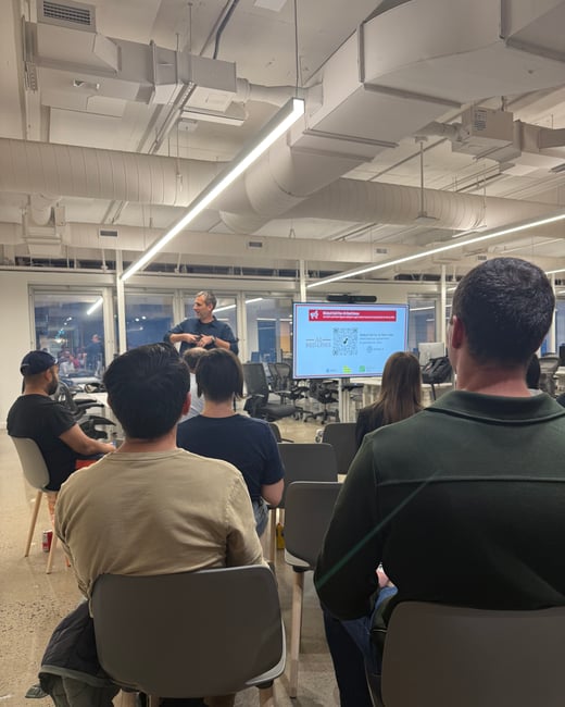 A photograph capturing an indoor presentation or lecture, showing a speaker standing in front of a screen addressing an audience seated in rows of chairs in a modern office space.