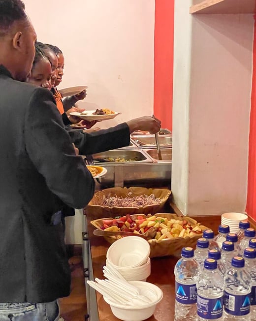 A group of people are standing in a line at a buffet, serving themselves from various food trays and bowls of salad and fruit.