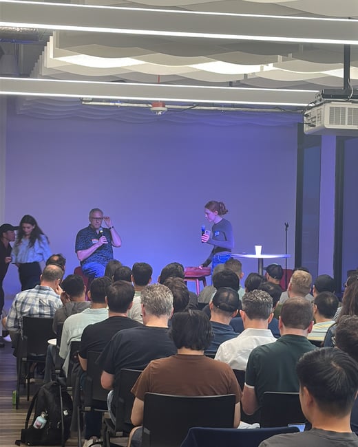 An indoor photograph showing a large audience seated in rows, facing two speakers who are addressing them from a stage area illuminated by purple ambient lighting.