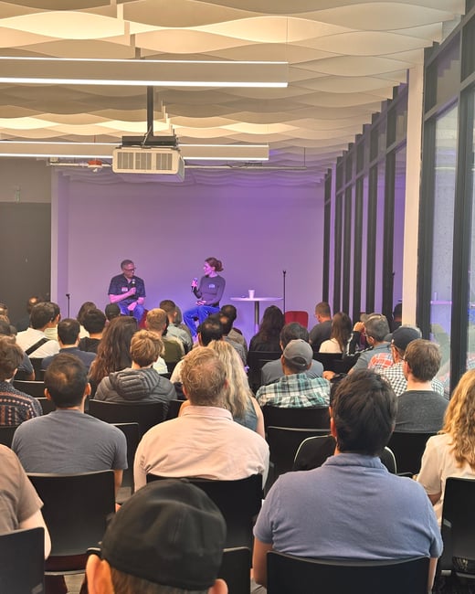 A photograph taken from the perspective of the audience, showing a large group of people seated and listening to two speakers on a stage illuminated with purple light.
