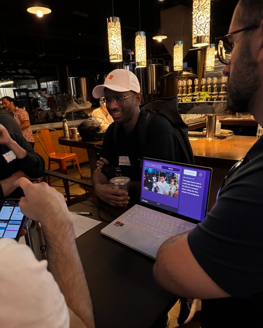 A candid photograph taken in a dimly lit indoor venue, likely a bar or brewery, showing a group of people gathered around a table, with a man in a white cap smiling while looking at a laptop displaying a text description.