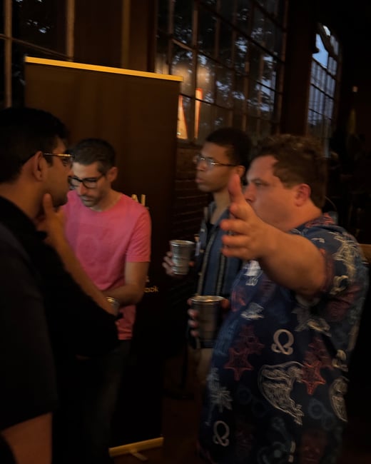 A dimly lit candid photograph captures four men socializing indoors at an event, possibly a networking gathering, with the man on the right gesturing while holding a metallic cup.