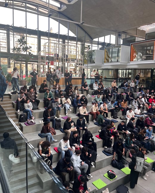 A photograph taken from an elevated perspective capturing a large group of young people sitting on tiered concrete steps in a brightly lit, modern indoor atrium, many of whom are looking at their mobile phones or chatting. The industrial-style building features a glass roof and large windows, suggesting a university or corporate gathering space.
