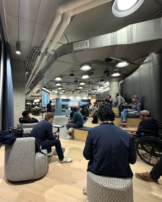 A group of people gathered in a modern, industrial-style indoor space, seemingly for a presentation or meeting. The room features tiered seating, exposed ceiling ducts, and a projection screen.