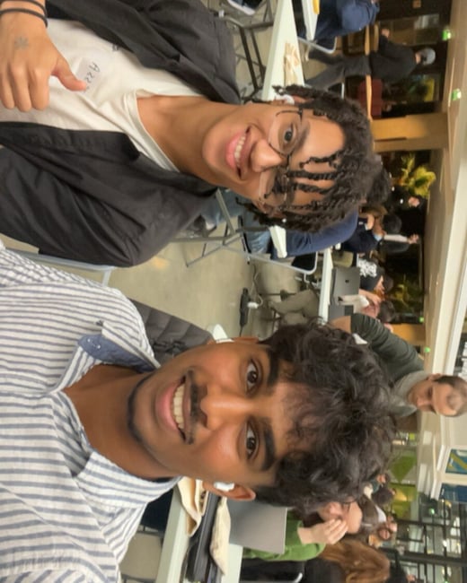 A close-up selfie featuring two young men smiling happily at the camera, taken indoors in a large, crowded event or workspace setting with many people seated at tables in the background.