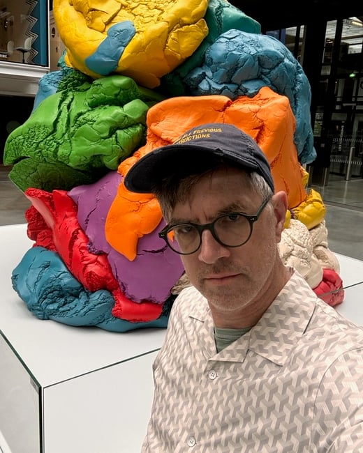 A man wearing glasses and a baseball cap takes a close-up selfie in an indoor setting, posing in front of a massive, brightly colored, abstract sculpture composed of textured, blob-like masses.