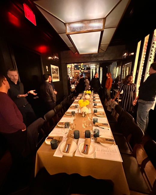 A photograph captures a group of people socializing around a long, formally set dining table in a dimly lit, upscale restaurant or private room with dark walls and a modern rectangular ceiling light.