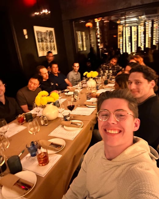 A close-up selfie captures a large group of people seated around a long dining table in a dimly lit, upscale restaurant setting. The person taking the photo, a young man wearing glasses, is smiling directly at the camera.