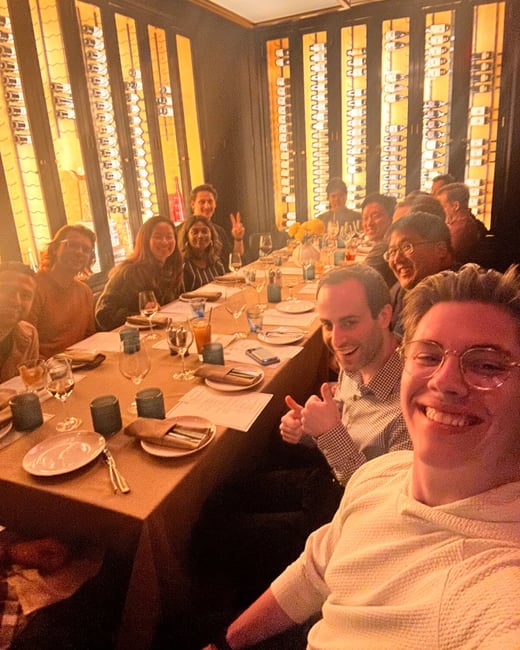 A group of approximately eleven people is gathered around a dining table in a warmly lit, upscale restaurant, smiling and posing for a group selfie. The background features dramatic, illuminated vertical wine racks built into the wall.