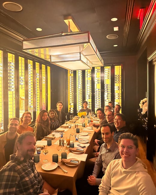 A group of approximately fourteen people is seated around a long dining table in a dimly lit, upscale restaurant or private room, posing for a group photo during a social dinner.
