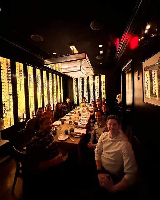A dimly lit photograph captures a large group of approximately fifteen people seated around a long dining table in an upscale restaurant or private room.