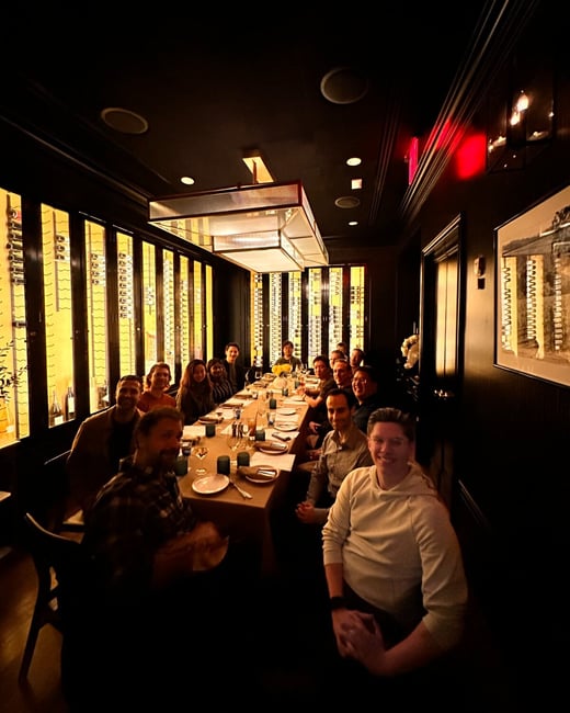 A large group of approximately seventeen people is gathered around a long dining table in a dimly lit, upscale private dining room with dark walls and a ceiling-mounted rectangular light fixture. The room features floor-to-ceiling illuminated wine racks along the left wall, providing warm background lighting.