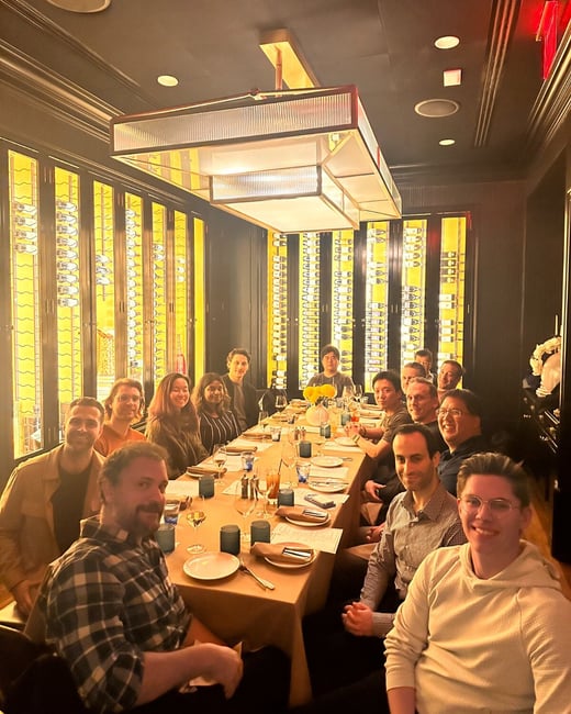 A group of approximately fifteen people is seated around a long dining table in a dimly lit, upscale restaurant setting, characterized by dark walls and illuminated wine racks in the background. Several individuals are smiling directly toward the camera, suggesting a social or business dinner gathering.