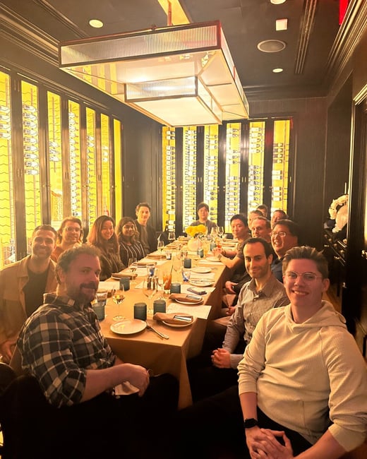 A photograph captures a large group of people seated around a long dining table in a dimly lit, upscale restaurant setting, characterized by dramatic yellow backlighting and a prominent overhead chandelier.